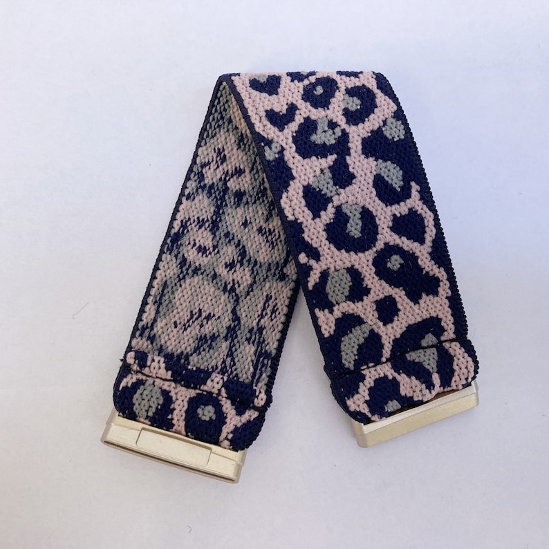 May include: A reversible elastic belt with a pink and blue leopard print on one side and a blue and pink floral print on the other side. The belt has gold metal buckles on both ends.
