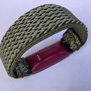 Op de afbeelding: Een geweven groen bandje met een roze fitness tracker eraan bevestigd. Het bandje is gemaakt van een zacht, flexibel materiaal en heeft een geweven patroon.