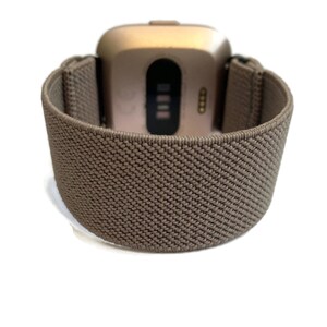 Op de afbeelding: Een bruin elastisch horlogebandje voor een smartwatch in goudkleur. Het bandje is gemaakt van geweven materiaal en heeft een textuur.
