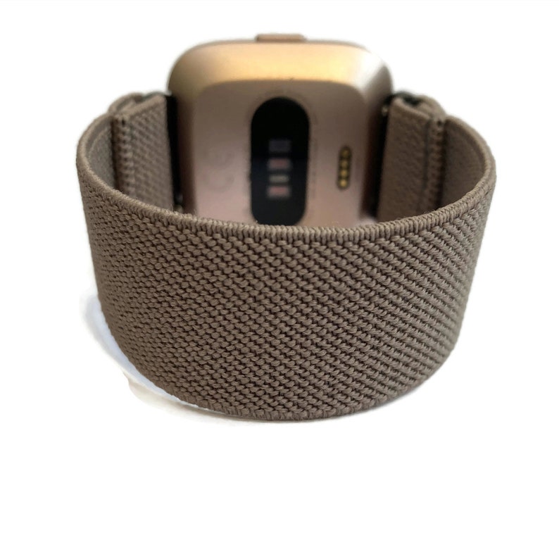 Op de afbeelding: Een bruin elastisch horlogebandje voor een smartwatch in goudkleur. Het bandje is gemaakt van geweven materiaal en heeft een textuur.