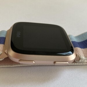 May include: A rose gold Fitbit smartwatch with a black screen and a pink, blue and white striped elastic band.
