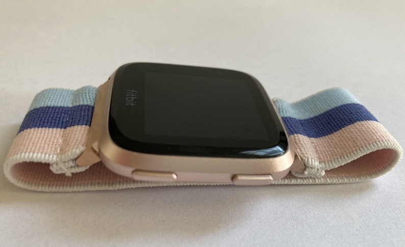 May include: A rose gold Fitbit smartwatch with a black screen and a pink, blue and white striped elastic band.