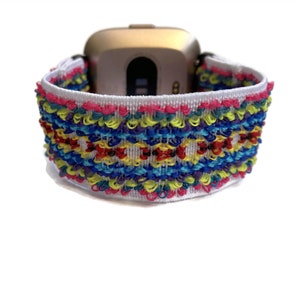 May include: A white elastic watch strap with a colourful embroidered pattern. The strap is designed for a smartwatch with a square face.
