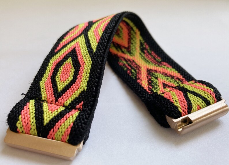 May include: A black elastic watch band with a geometric pattern in pink and green. The band has a gold metal clasp.