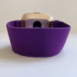 Pu&ograve; includere: Uno smartwatch color oro con un cinturino a coste viola scuro. Il cinturino &egrave; realizzato in un tessuto strutturato ed &egrave; progettato per adattarsi al polso. Il quadrante dell'orologio &egrave; visibile.