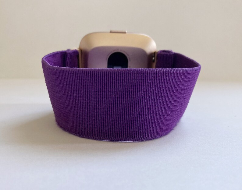Pu&ograve; includere: Uno smartwatch color oro con un cinturino a coste viola scuro. Il cinturino &egrave; realizzato in un tessuto strutturato ed &egrave; progettato per adattarsi al polso. Il quadrante dell'orologio &egrave; visibile.