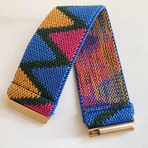 May include: A colorful woven elastic watch band with a geometric pattern in shades of blue, green, pink, and orange. The band has gold metal clasps on each end.
