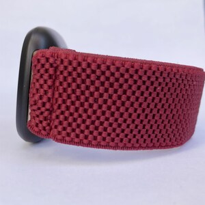 May include: A close-up of a red watch band with a black watch face. The band is made of a soft, breathable material with a textured pattern.