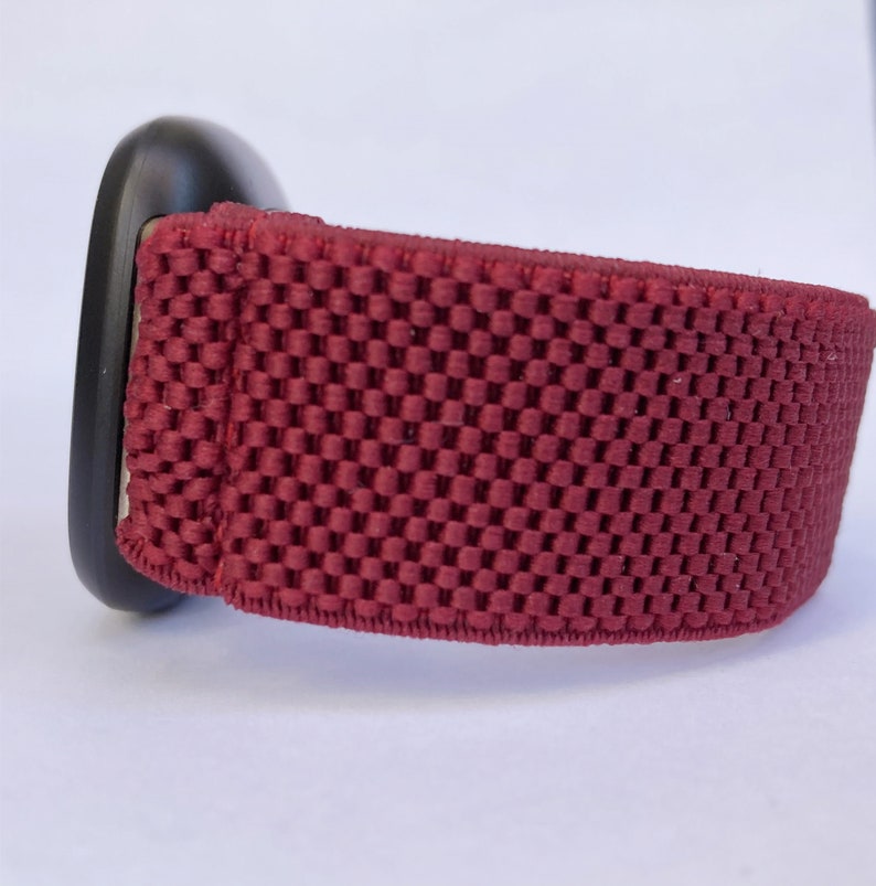 May include: A close-up of a red watch band with a black watch face. The band is made of a soft, breathable material with a textured pattern.