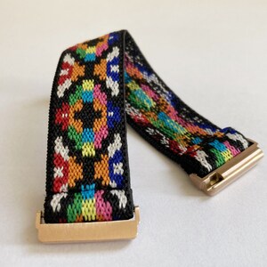 May include: A black elastic watch band with a colorful geometric pattern and gold metal clasps.
