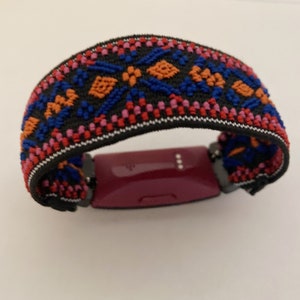 Op de afbeelding: Een zwart en kleurrijk bandje met patroon voor een fitness tracker. Het bandje heeft een geometrisch ontwerp met rode, blauwe, oranje en witte accenten.