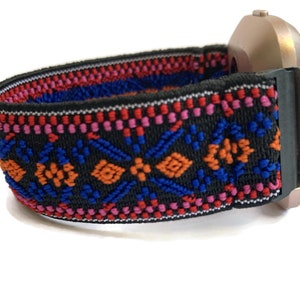 Op de afbeelding: Een zwart horlogebandje met een kleurrijk geometrisch patroon in rood, oranje, blauw en roze. Het bandje is gemaakt van elastisch materiaal en heeft een zilveren horlogekast.