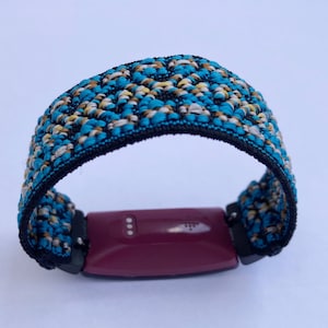 Op de afbeelding: Een geweven band in blauw, wit en goud voor een fitness tracker. De band is bevestigd aan een bordeauxrode fitness tracker.