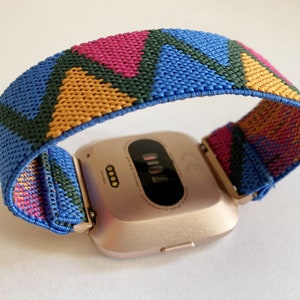 May include: A rose gold smartwatch with a colorful geometric patterned band. The band is made of woven fabric and features blue, green, pink, and yellow triangles.
