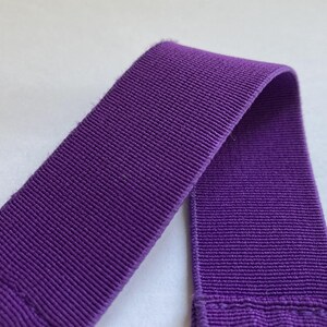 Pu&ograve; includere: Primo piano di un pezzo di elastico viola a coste. L'elastico &egrave; di un colore viola intenso e vibrante e ha un aspetto strutturato e tessuto. Il materiale &egrave; ripiegato su se stesso, mostrando la trama e la costruzione del tessuto.