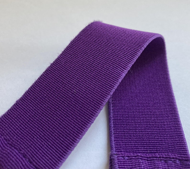 Pu&ograve; includere: Primo piano di un pezzo di elastico viola a coste. L'elastico &egrave; di un colore viola intenso e vibrante e ha un aspetto strutturato e tessuto. Il materiale &egrave; ripiegato su se stesso, mostrando la trama e la costruzione del tessuto.