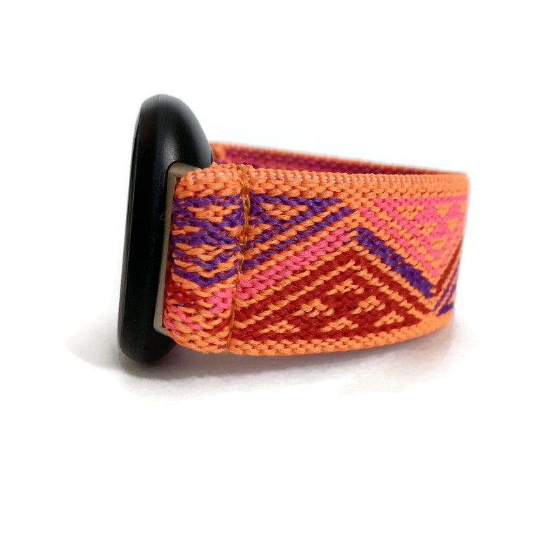 May include: A close-up of a bright orange watch band with a geometric pattern in shades of pink and purple. The band is made of woven fabric and is attached to a black watch face.