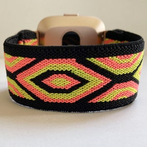 May include: Black elastic watch band with a geometric pattern in pink and green. The band is designed to fit a smartwatch.