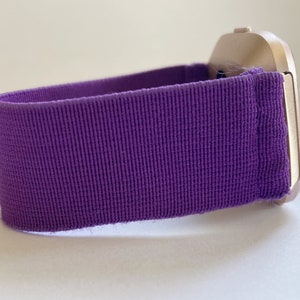 Pu&ograve; includere: Primo piano di uno smartwatch con cinturino in tessuto viola. La cassa dell'orologio &egrave; color oro chiaro. Il cinturino &egrave; viola a tinta unita e strutturato. L'orologio &egrave; su una superficie bianca.