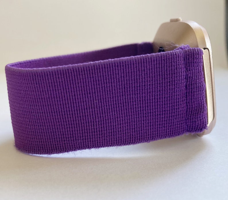 Pu&ograve; includere: Primo piano di uno smartwatch con cinturino in tessuto viola. La cassa dell'orologio &egrave; color oro chiaro. Il cinturino &egrave; viola a tinta unita e strutturato. L'orologio &egrave; su una superficie bianca.