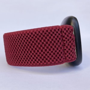 May include: A close-up of a burgundy red watch band with a woven texture. The band is attached to a black watch face.