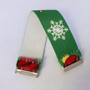 Puede incluir: Una pulsera el&aacute;stica verde y blanca con un dise&ntilde;o de copo de nieve y adornos navide&ntilde;os. La pulsera tiene cierres met&aacute;licos plateados en cada extremo.