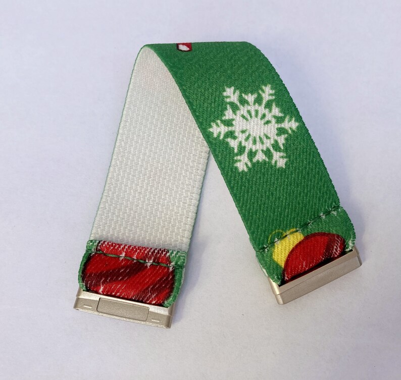 Puede incluir: Una pulsera el&aacute;stica verde y blanca con un dise&ntilde;o de copo de nieve y adornos navide&ntilde;os. La pulsera tiene cierres met&aacute;licos plateados en cada extremo.