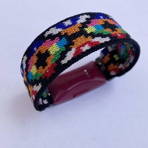 May include: A colourful woven band with a black background and a pink fitness tracker attached.