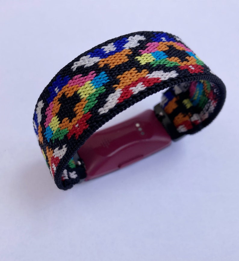 May include: A colourful woven band with a black background and a pink fitness tracker attached.