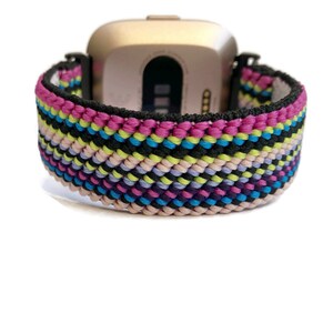 May include: A colourful woven watch band with pink, blue, green, black and white stripes. The band is made of a soft, flexible material and is compatible with Fitbit Versa smartwatches.