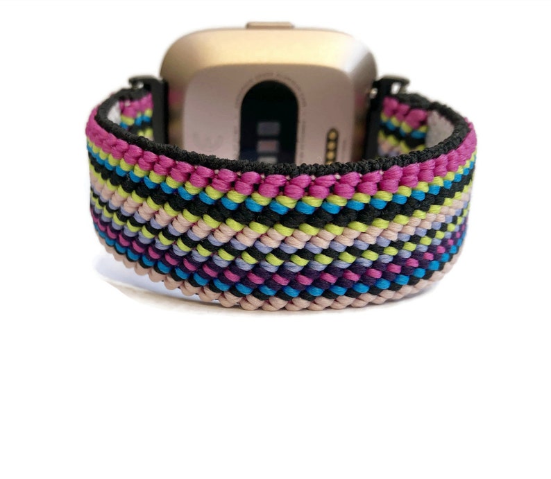 May include: A colourful woven watch band with pink, blue, green, black and white stripes. The band is made of a soft, flexible material and is compatible with Fitbit Versa smartwatches.