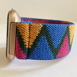 May include: A gold-colored smartwatch with a colorful geometric patterned band. The band is made of a woven fabric with pink, blue, green, and yellow stripes.
