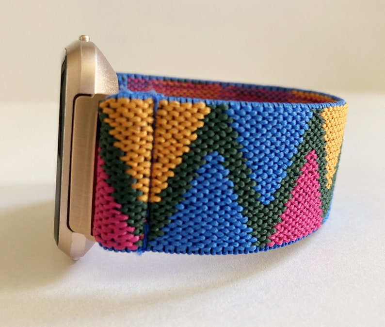 May include: A gold-colored smartwatch with a colorful geometric patterned band. The band is made of a woven fabric with pink, blue, green, and yellow stripes.