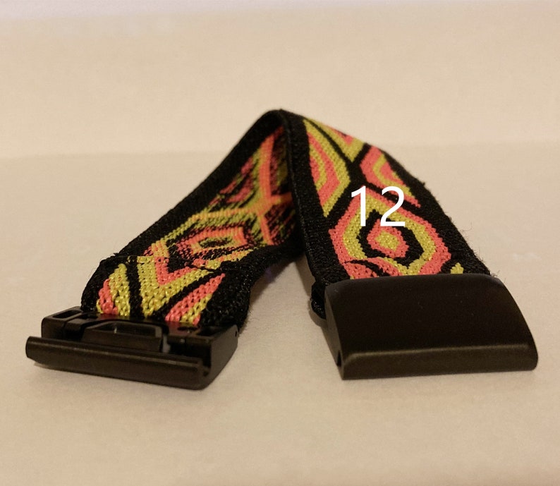 Op de afbeelding: Een zwart en geel gepatroneerd horlogebandje met een zwarte gesp. Het bandje heeft een roze en geel geometrisch ontwerp. Het getal "12" is op het bandje gedrukt.