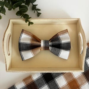 May include: A plaid bow tie in brown, black, and white, resting on a wooden tray. The bow tie has a classic shape and is made of a fabric with a large check pattern. A matching fabric is visible below the tray.