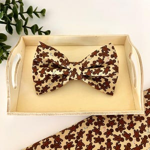 May include: A brown and tan bow tie with a gingerbread man pattern, resting on a wooden tray. The bow tie is made of fabric and features a repeating design of gingerbread men and candy canes. A matching fabric piece is visible.
