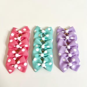 May include: Three stacks of polka dot hair bows in pink, teal, and lavender. Each bow has a white center and white polka dots. The bows are arranged on a white background.