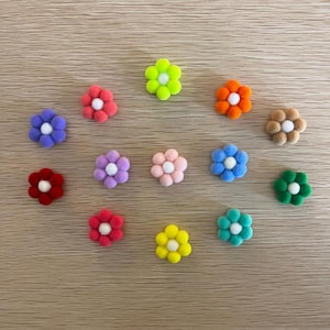 May include: A collection of colorful, small, felt flower embellishments. The flowers are in various colors, including red, pink, purple, blue, green, yellow, orange, and brown, each with a white center. They are arranged on a light brown wooden surface.