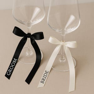 Peut inclure: Deux verres à vin transparents ornés de rubans décoratifs. L'un est orné d'un ruban noir avec le mot « GROOM » en blanc, l'autre d'un ruban ivoire avec « BRIDE » en noir. Les rubans sont noués autour des pieds.