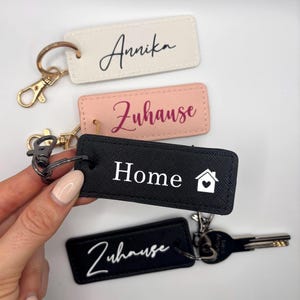 Könnte beinhalten: Vier Schlüsselanhänger mit verschiedenen Designs. Der erste Schlüsselanhänger ist weiß mit schwarzem kursivem Text, der "Annika" sagt. Der zweite Schlüsselanhänger ist rosa mit burgunderfarbenem kursivem Text, der "Zuhause" sagt. Der dritte Schlüsselanhänger ist schwarz mit weißem Text, der "Home" sagt und einem weißen Haussymbol mit einem roten Herzen. Der vierte Schlüsselanhänger ist schwarz mit weißem kursivem Text, der "Zuhause" sagt.
