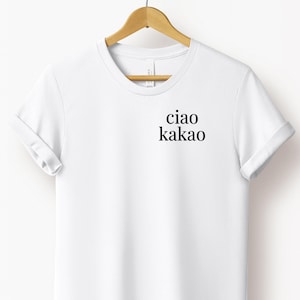 Könnte beinhalten: Weißes T-Shirt mit den Worten "ciao kakao" in Schwarz gedruckt. Das Shirt hängt an einem Holzbügel. Die Ärmel sind umgeschlagen.