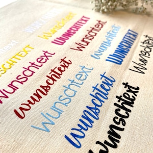Könnte beinhalten: Nahaufnahme einer weißen Stofftasche mit dem Wort "Wunschtext" in verschiedenen Farben gestickt, darunter Gelb, Rot, Hellblau, Blau und Schwarz.