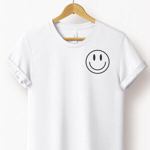 Könnte beinhalten: Weißes T-Shirt mit einem schwarzen Smiley-Gesicht auf der Brust. Das kurzärmelige T-Shirt hängt an einem Holzbügel. Das T-Shirt besteht aus einem weichen Material.