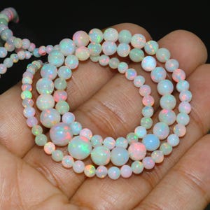 May include: Close-up of multiple strands of opal beads. The beads are predominantly white with flashes of pink, green, and blue, creating an iridescent effect. The beads are round and come in different sizes.