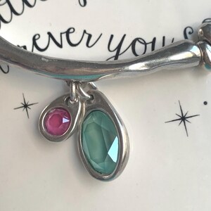 May include: Silver bracelet with two gemstone charms. One charm is a small, round pink gemstone, and the other is a larger, oval-shaped green gemstone. The bracelet is set against a white background with the words "little sparkle" and "ever you" in black script.