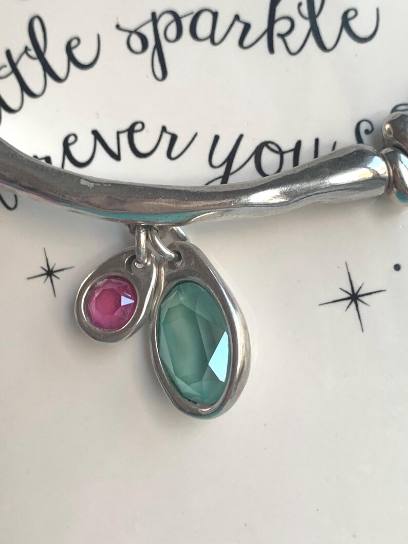 May include: Silver bracelet with two gemstone charms. One charm is a small, round pink gemstone, and the other is a larger, oval-shaped green gemstone. The bracelet is set against a white background with the words "little sparkle" and "ever you" in black script.