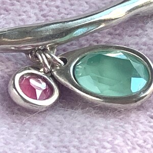 May include: A silver bracelet with two gemstone pendants. One pendant is an oval-shaped, light green gemstone, and the other is a smaller, round, pink gemstone. The bracelet is set against a soft, light purple background.