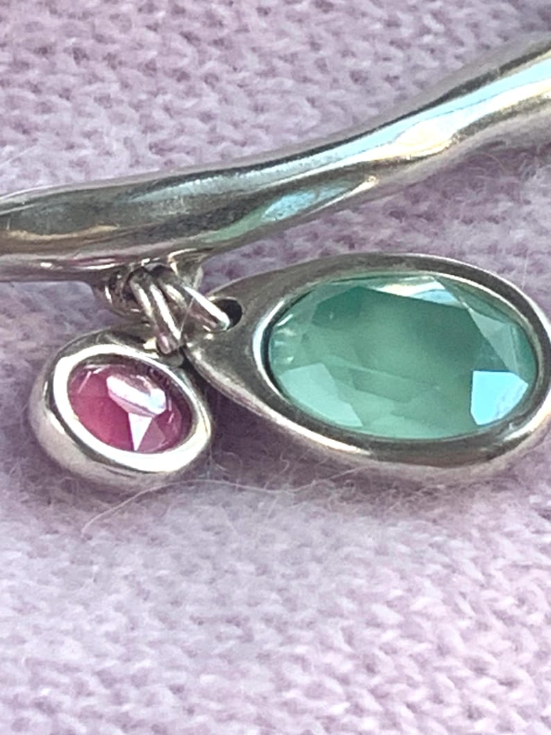 May include: A silver bracelet with two gemstone pendants. One pendant is an oval-shaped, light green gemstone, and the other is a smaller, round, pink gemstone. The bracelet is set against a soft, light purple background.
