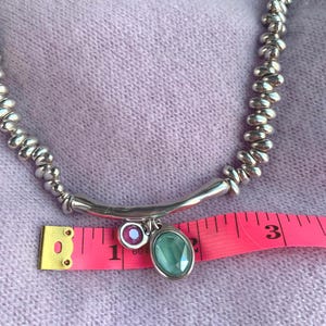 May include: Silver-toned necklace with a beaded chain and a curved bar pendant. The pendant features a small, round, purple gemstone and a larger, oval, teal gemstone. The necklace is measured with a pink measuring tape.