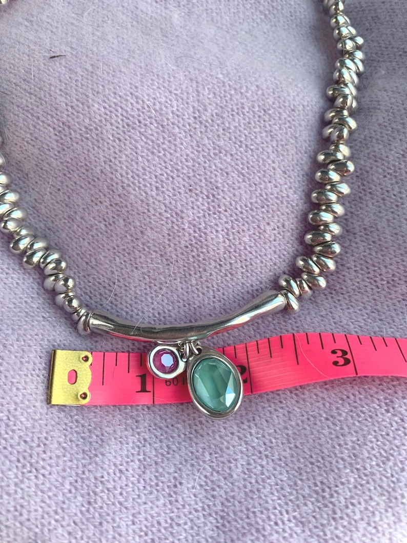 May include: Silver-toned necklace with a beaded chain and a curved bar pendant. The pendant features a small, round, purple gemstone and a larger, oval, teal gemstone. The necklace is measured with a pink measuring tape.